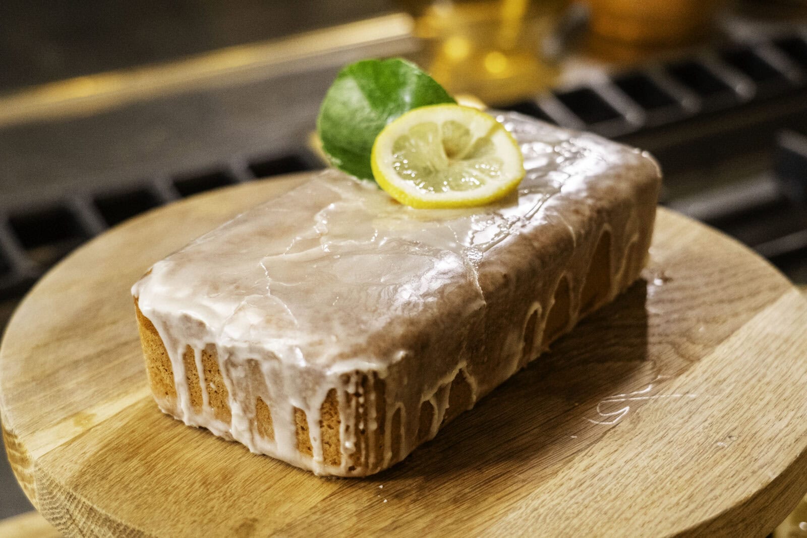 A glazed loaf cake topped with a lemon slice and green leaf sits on a wooden board.