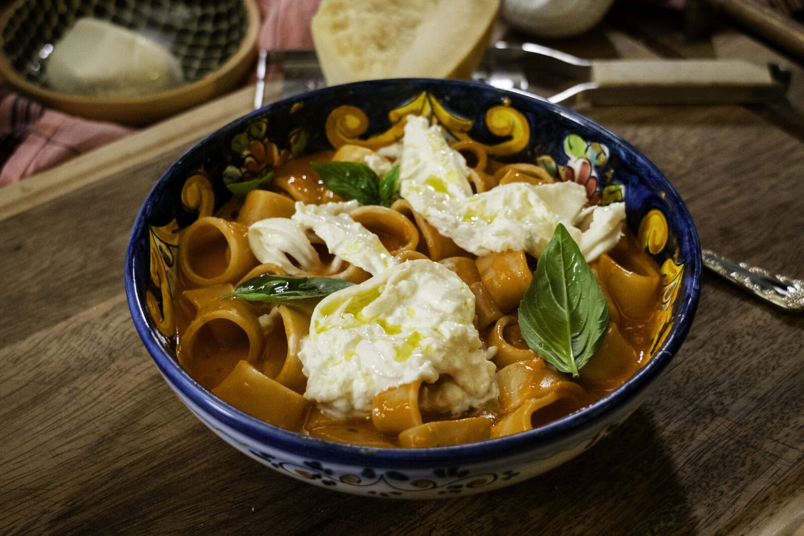 A bowl of pasta in tomato sauce topped with burrata cheese and fresh basil sits on a wooden surface beside a slice of bread.