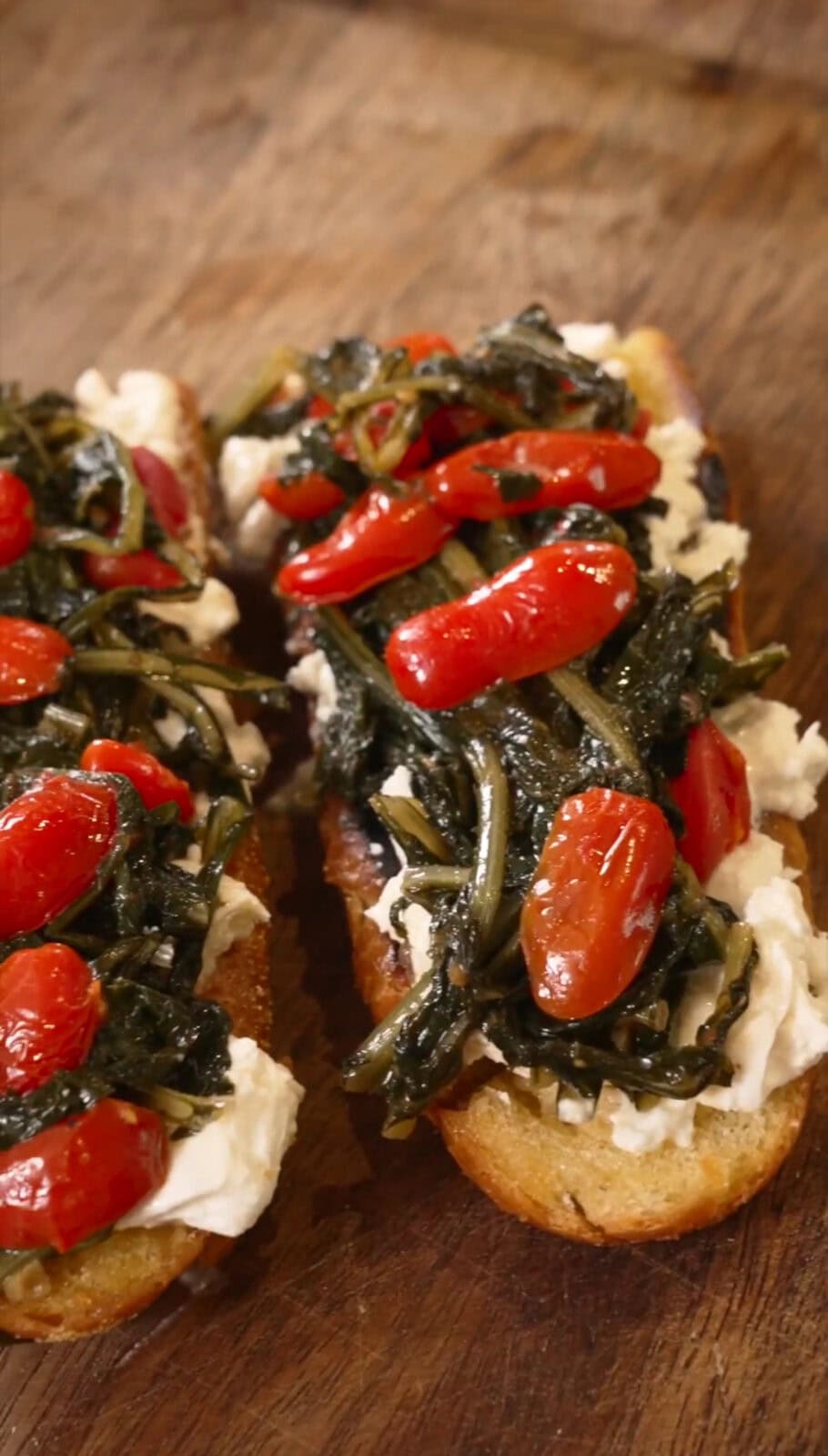 Two slices of toast topped with cream cheese, sautéed greens, and cherry tomatoes.