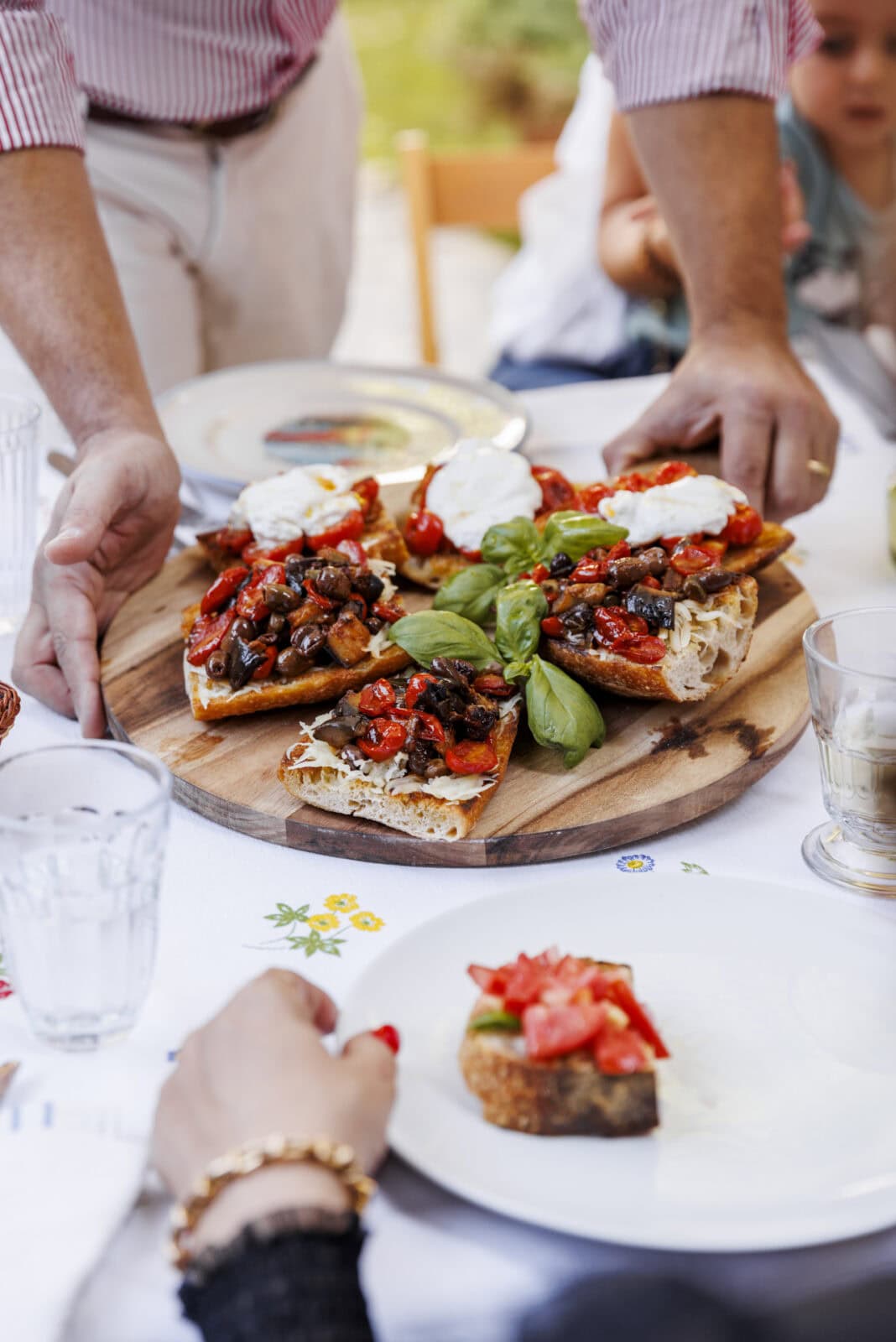 Hands placing a large wooden board with bruschetta topped with tomatoes, basil, and mozzarella on a table. A person reaches for a slice.