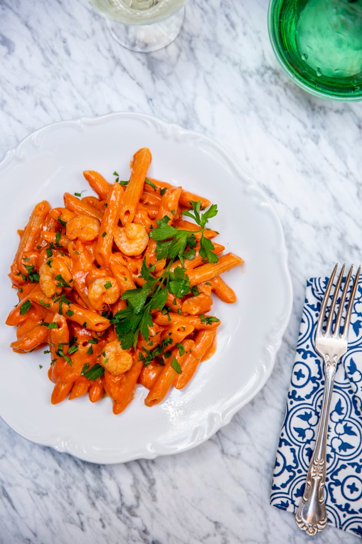 Plate of creamy penne pasta with shrimp, garnished with parsley. Placed on a marble surface near a patterned napkin and fork.