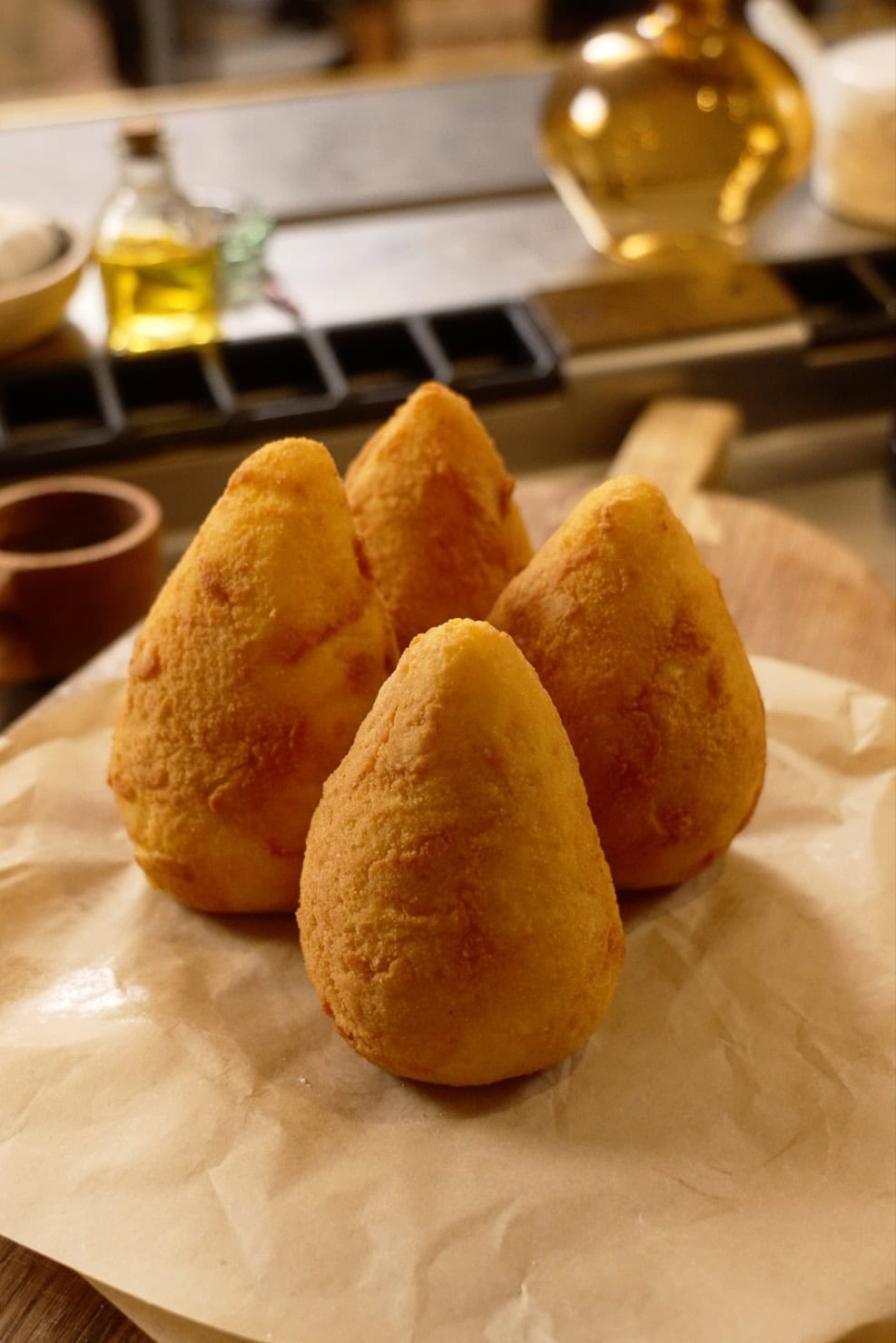 Four golden, cone-shaped fried rice balls sit on parchment paper, placed on a wooden board in a kitchen setting.