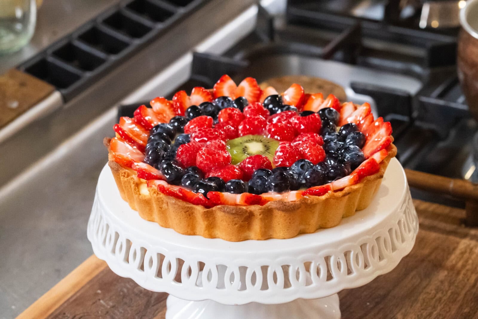Fruit tart with strawberries, blueberries, raspberries, and kiwi on a white stand, near a gas stove.