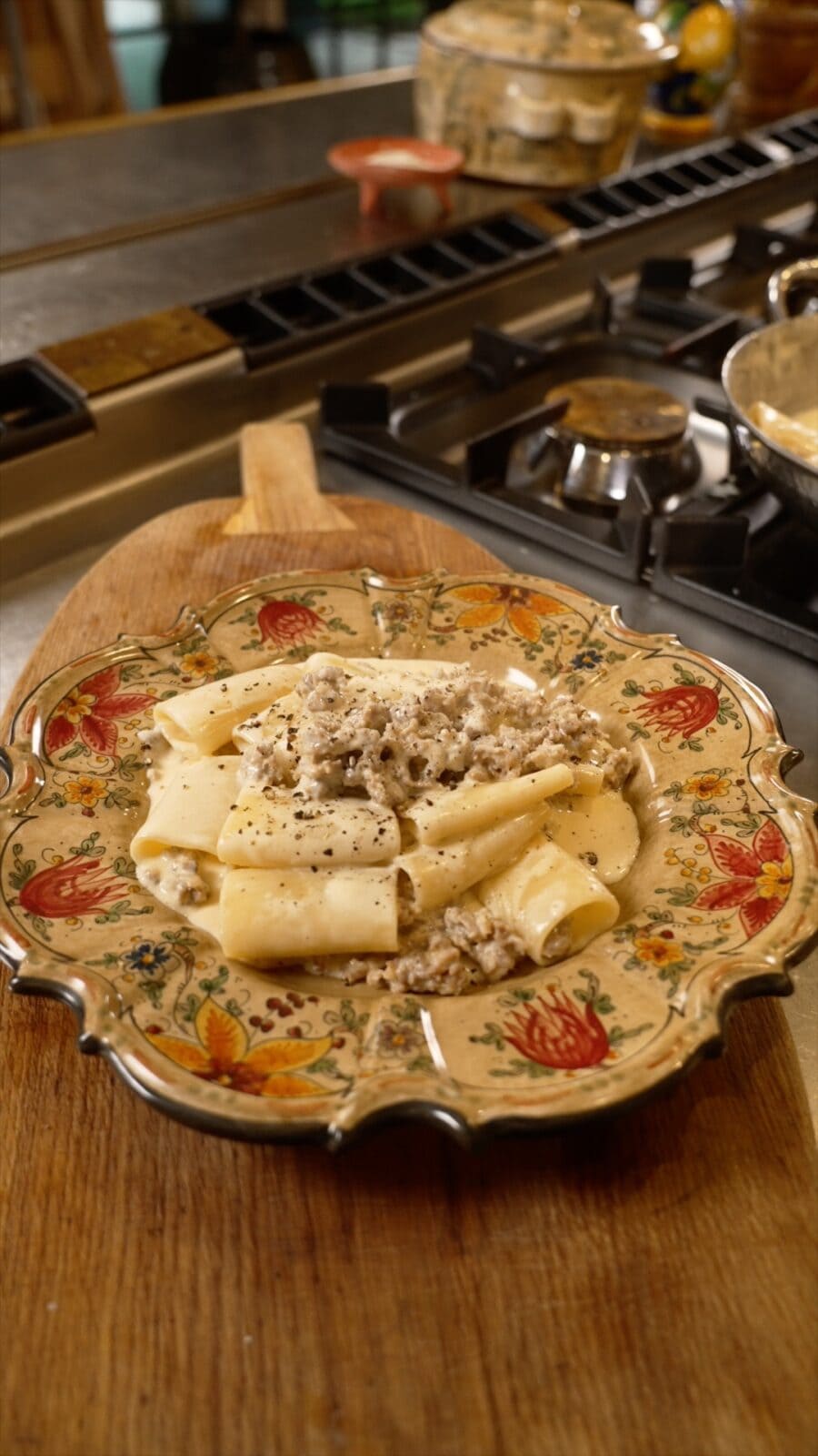A decorative plate of pasta topped with a creamy meat sauce sits on a wooden cutting board near a gas stove in a kitchen.