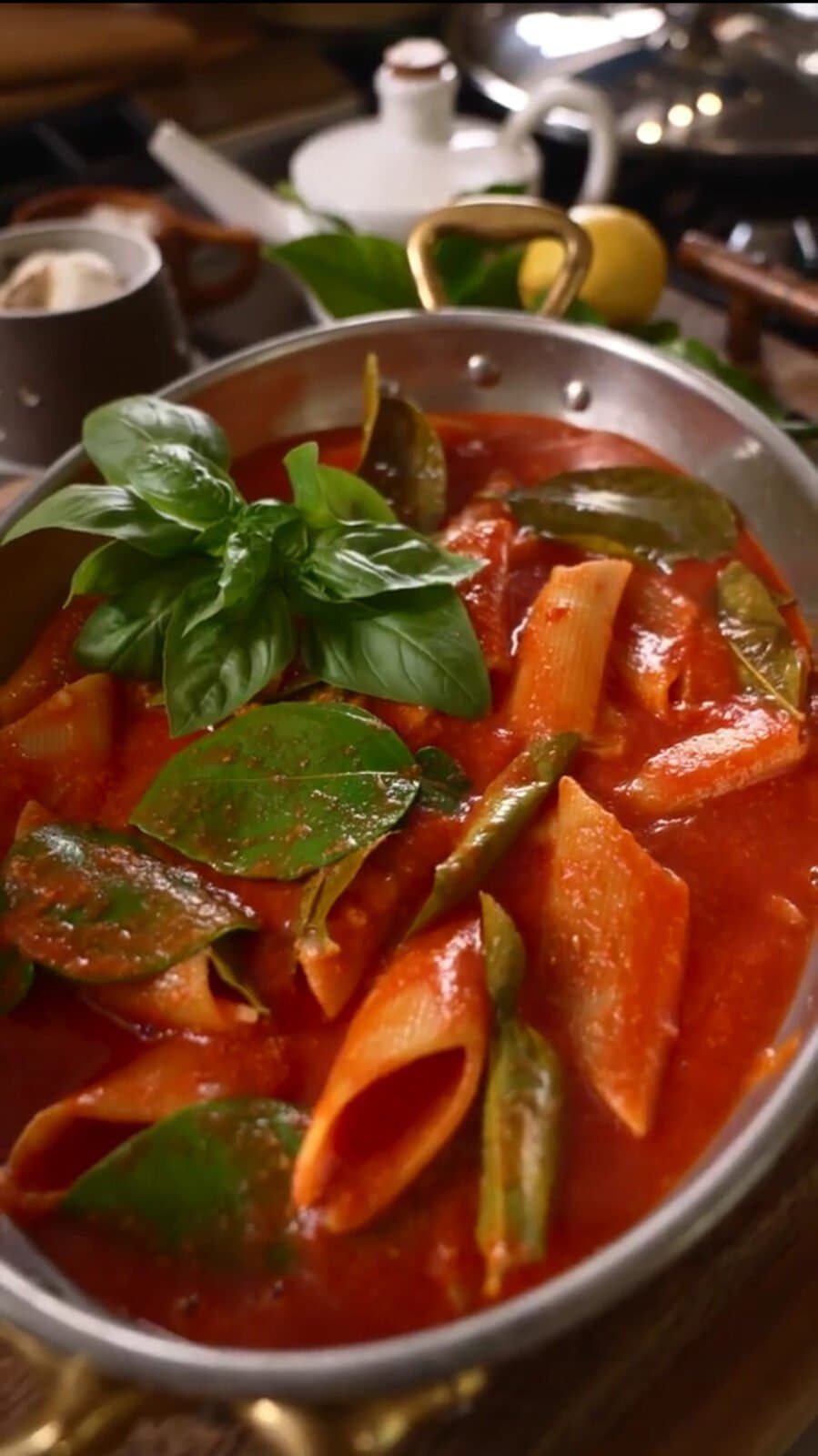 A pot of penne pasta in tomato sauce with basil leaves and vegetables, next to a lemon and a teacup on a wooden table.