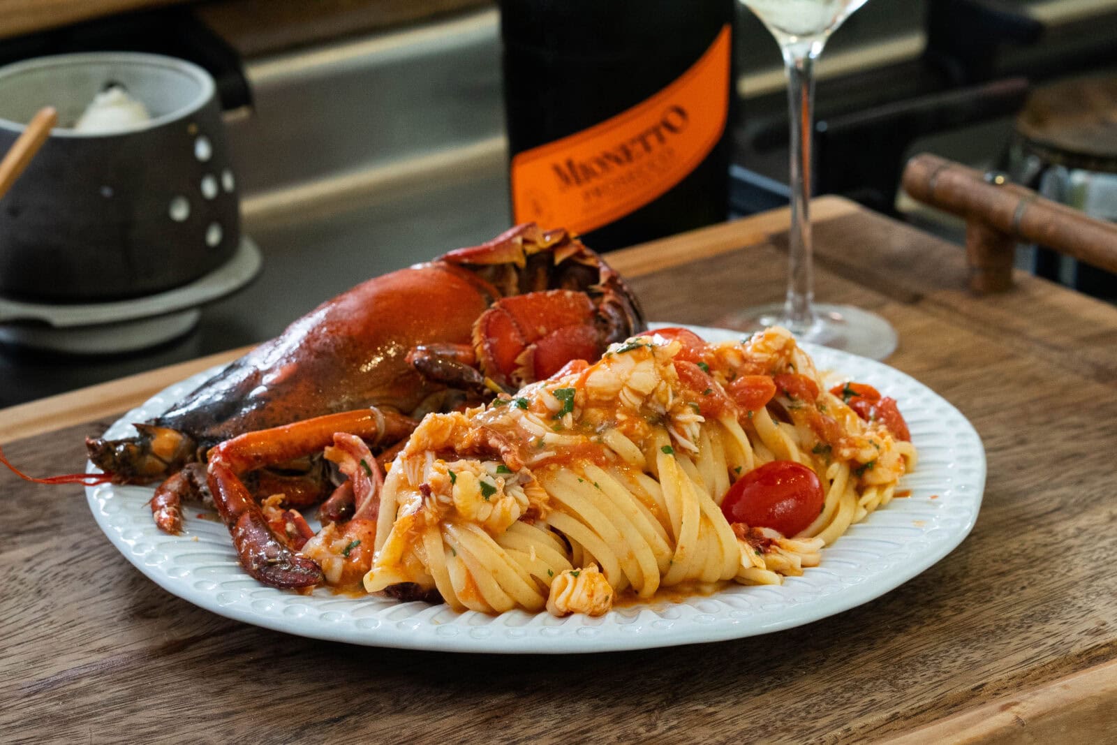 A plate of pasta with cherry tomatoes and lobster in a tomato-based sauce, served with a bottle of Mionetto and a wine glass on a wooden surface.