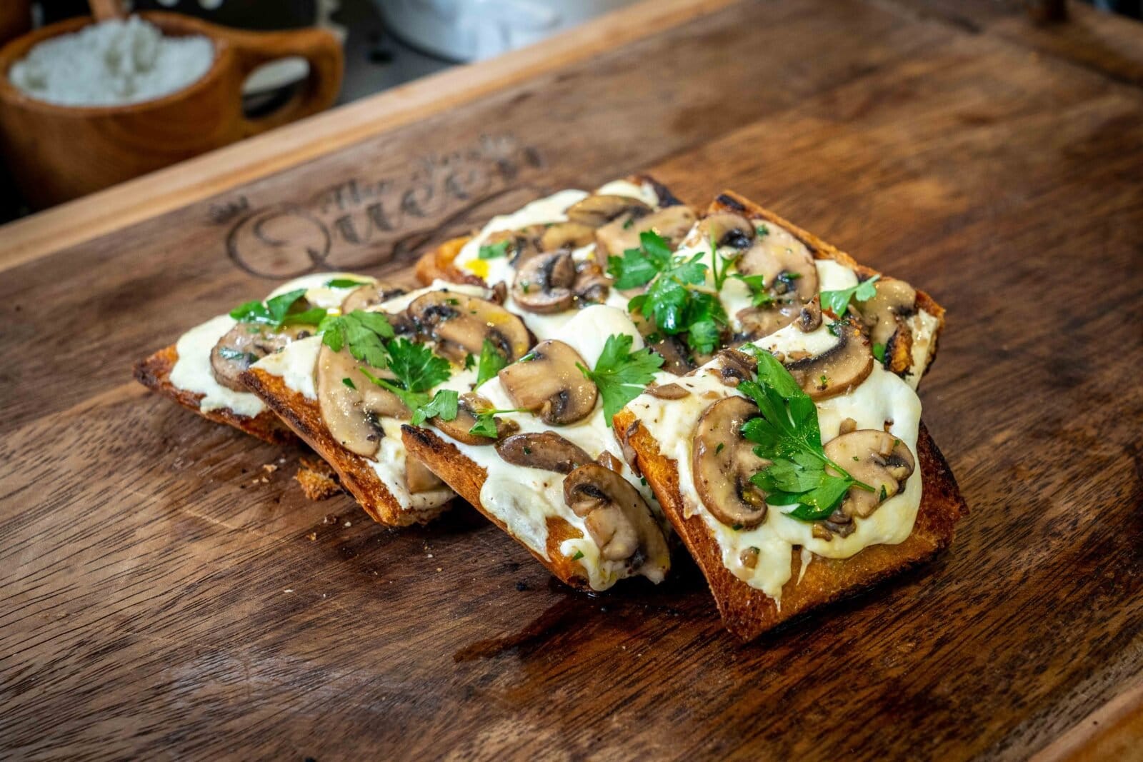 Four slices of toasted bread topped with melted cheese, sautéed mushrooms, and parsley on a wooden board.