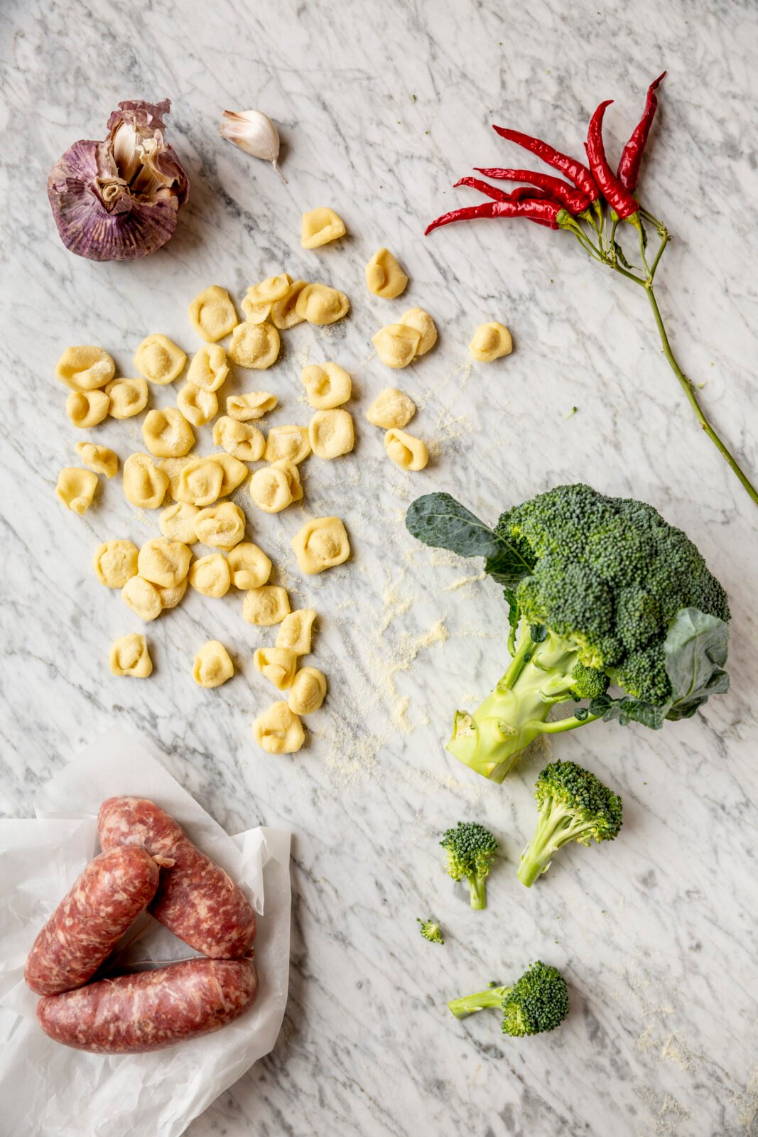 Pasta, broccoli, sausages, garlic, and red chili peppers are arranged on a marble surface.