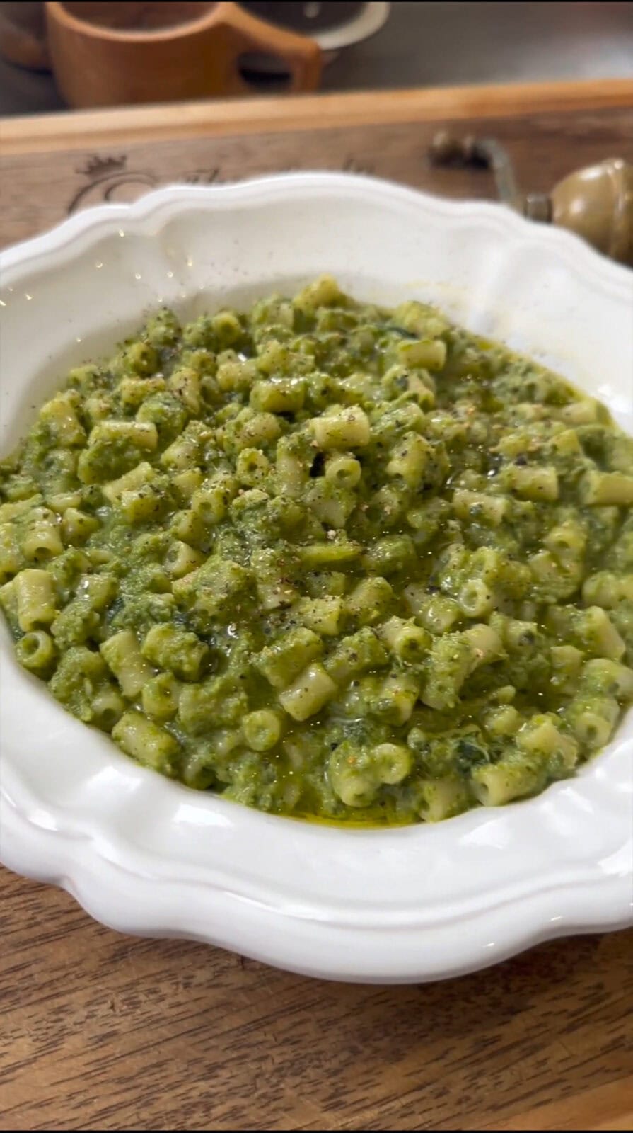 A white bowl filled with pasta mixed in a green pesto sauce, garnished with black pepper and olive oil, placed on a wooden table.