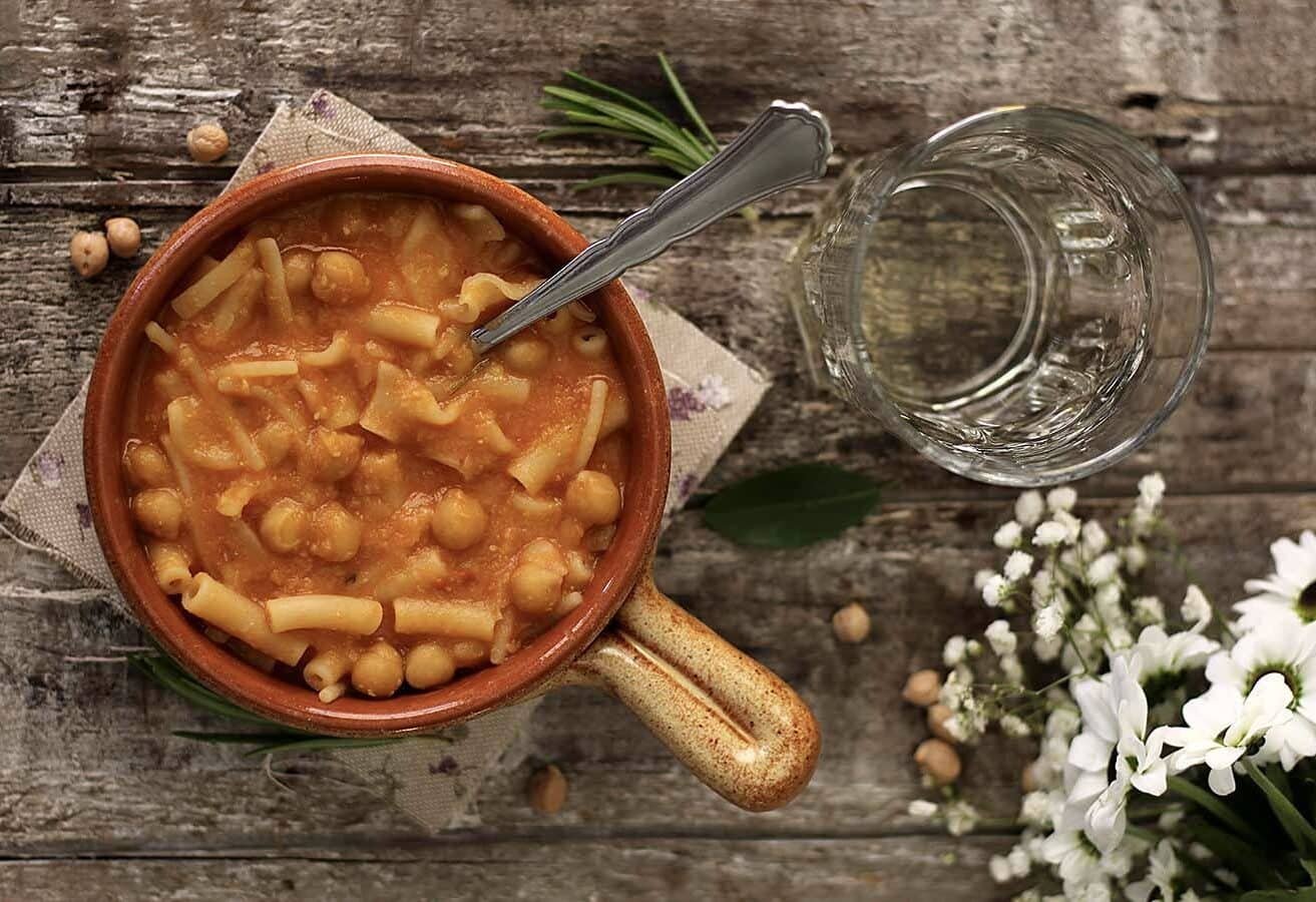 A bowl of pasta with chickpeas and tomato sauce on a rustic wooden surface. A spoon rests in the bowl. Nearby are a glass of water, white flowers, rosemary, and chickpeas.