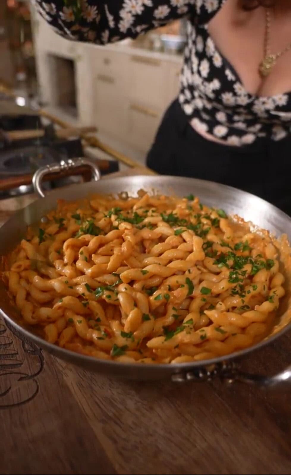 A skillet filled with cooked pasta in a creamy sauce, garnished with chopped parsley. A person in a floral top is visible in the background.