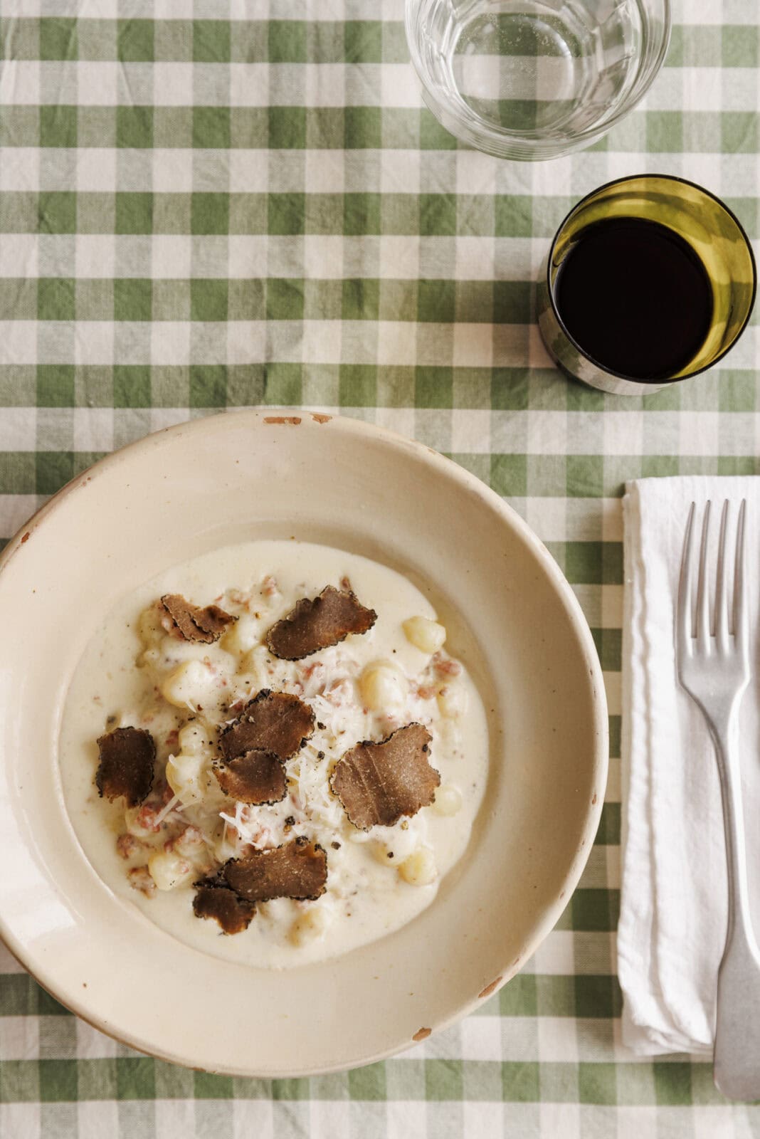 A bowl of creamy gnocchi topped with truffle slices, set on a green and white checkered tablecloth. A glass of water, a glass of red wine, a fork, and a knife are beside it.