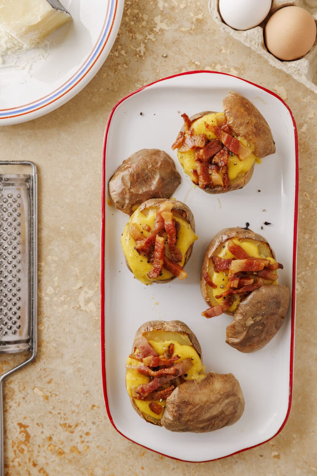 Four baked potatoes topped with melted cheese and bacon pieces on a white rectangular plate. Nearby are eggs, a cheese grater, and a block of cheese on a plate.