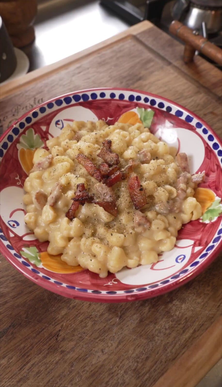 A colorful bowl filled with creamy macaroni and cheese topped with crispy bacon pieces and a sprinkle of black pepper, placed on a wooden surface.