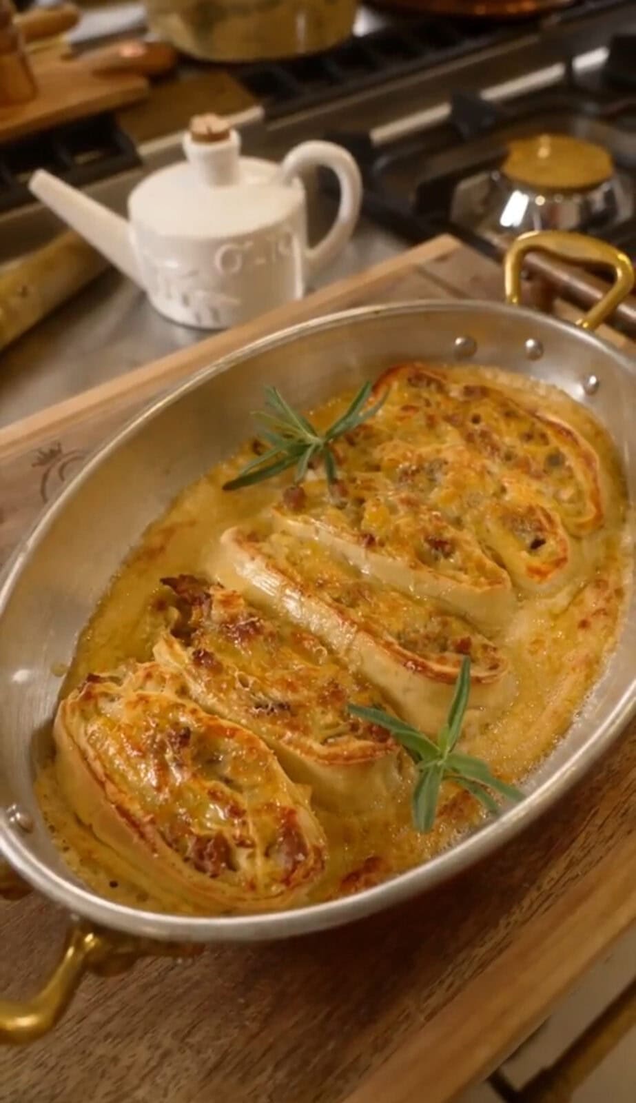 Oval dish with baked stuffed pasta rolls, garnished with rosemary sprigs, on a wooden board. Kettle visible in background.