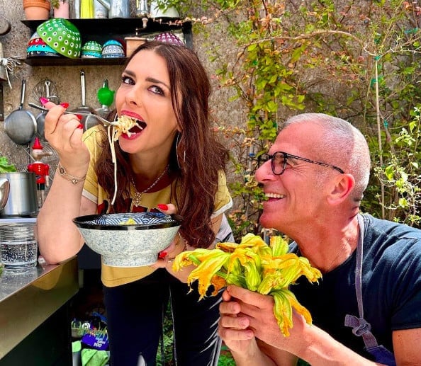 A woman eats pasta from a bowl with a fork while a man next to her holds fresh zucchini flowers and smiles; they are in an outdoor kitchen setting.