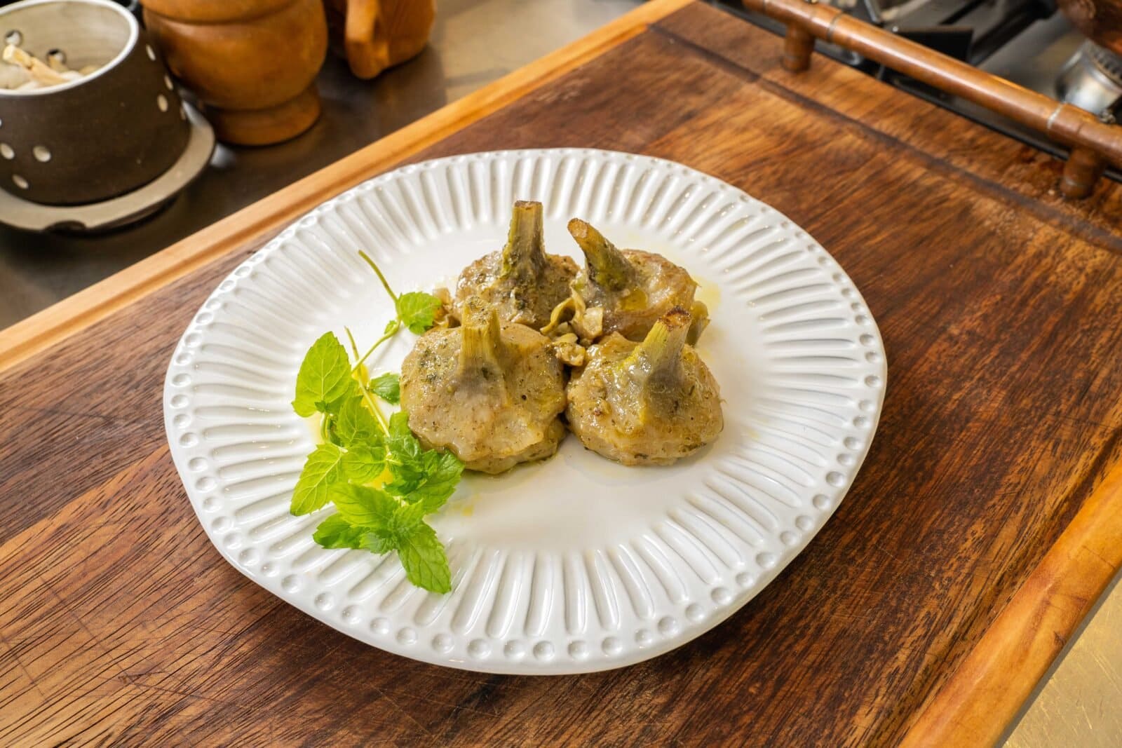 Three cooked artichokes with a garnish of mint leaves on a white plate, placed on a wooden surface.