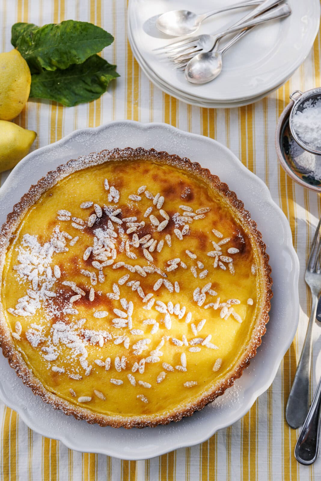 A lemon tart topped with pine nuts and powdered sugar sits on a white plate, surrounded by lemons, mint leaves, cutlery, and stacked plates on a striped tablecloth.