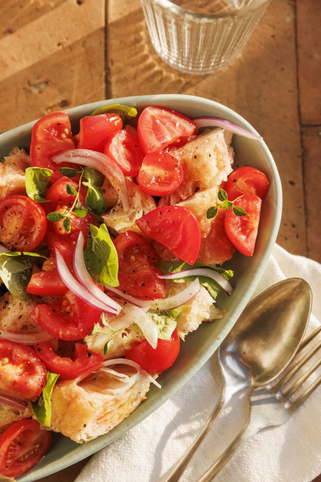 Oval dish with tomato, onion, basil, and bread salad on a napkin beside a spoon, fork, and glass of water on a sunlit table.