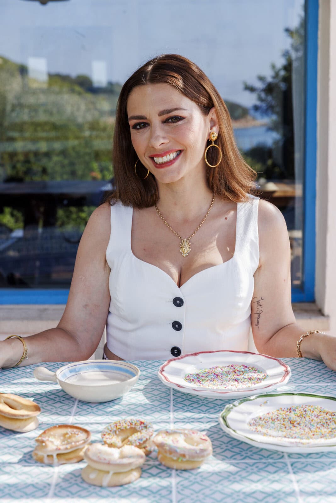 A woman in a white top sits at a table with plates of bagels, bowls of milk, and colorful sprinkles in front of a window on a sunny day.