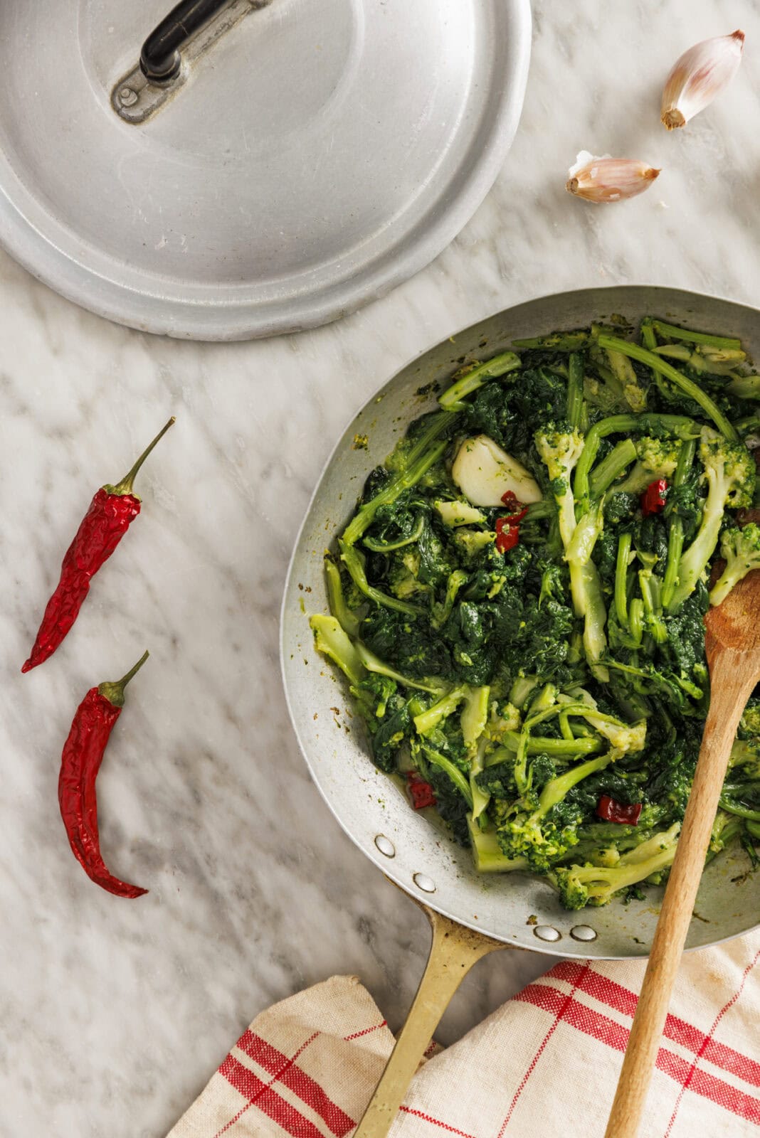 A pan of cooked leafy greens with chopped red chili peppers sits on a marble countertop, surrounded by a pot lid, two garlic cloves, two dried red chilies, and a red-striped towel.