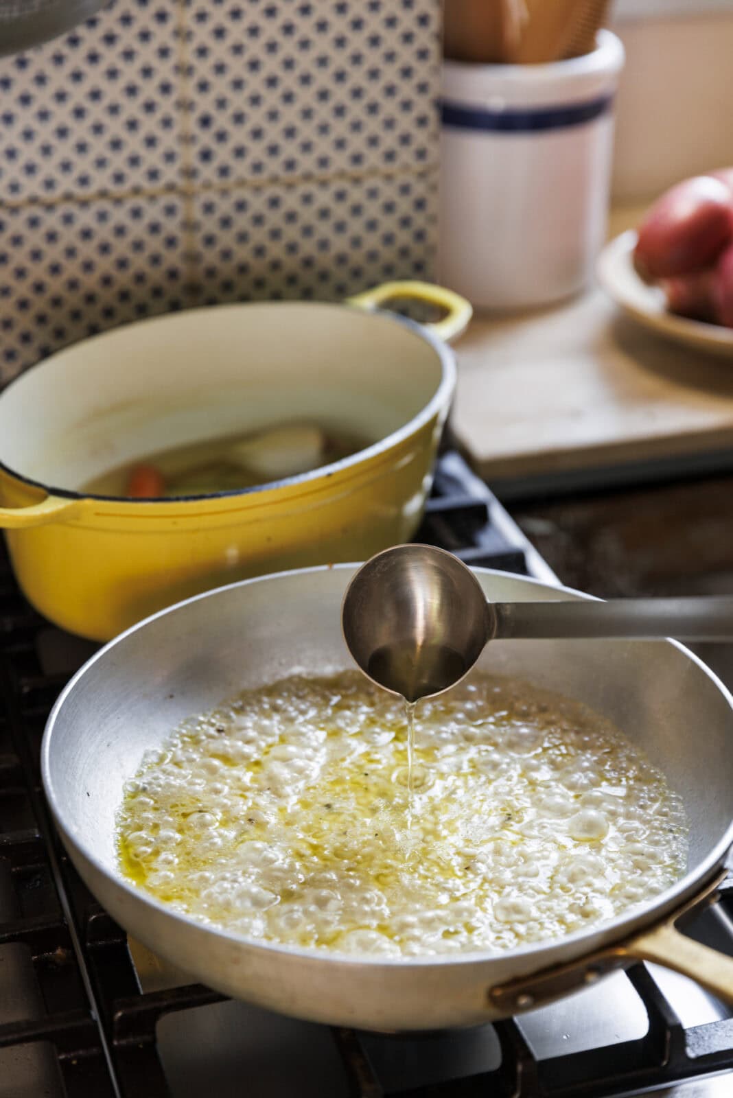 A metal ladle pours liquid into a skillet of bubbling oil on a stovetop, with a yellow Dutch oven and a plate of onions in the background.