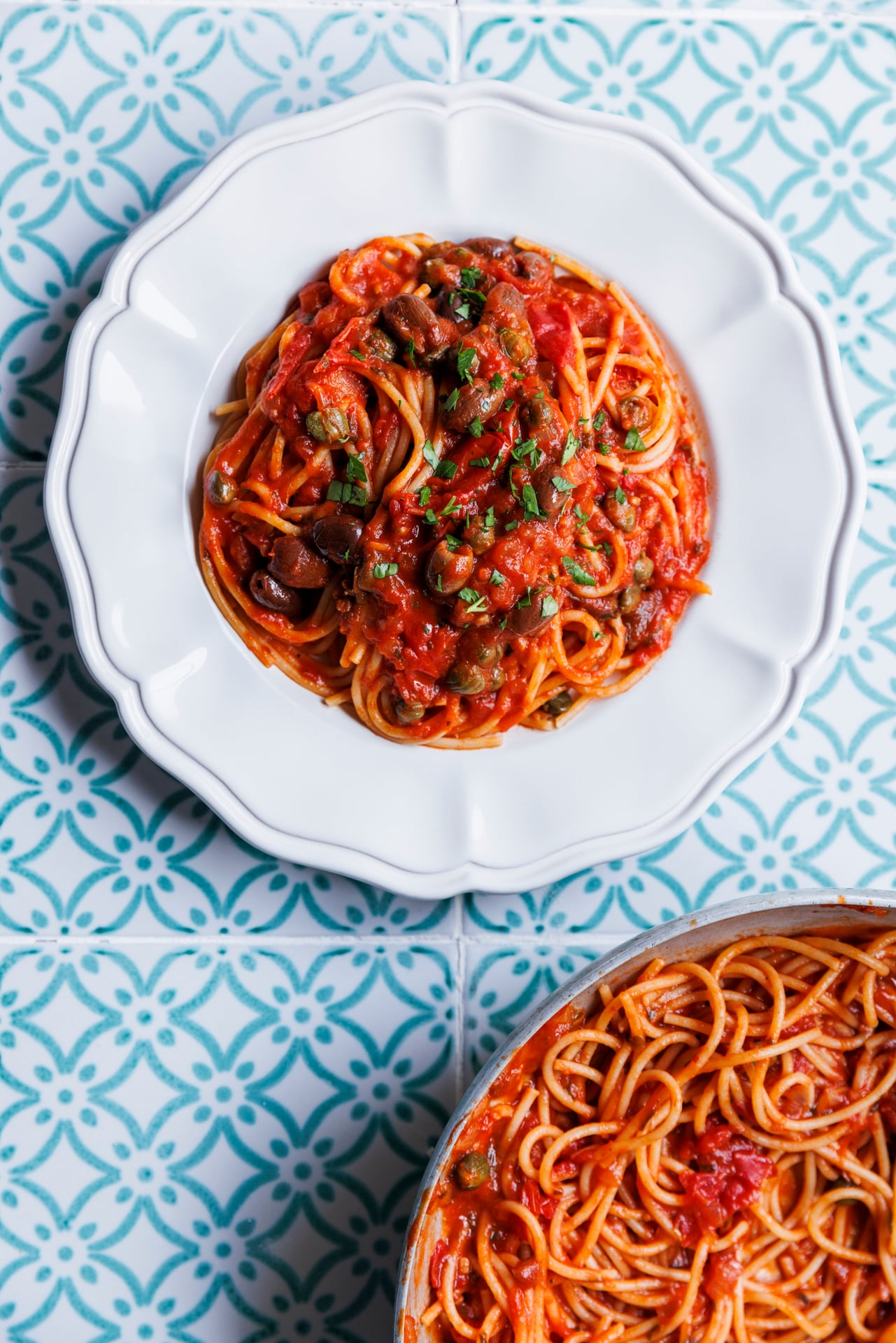 A plate of spaghetti with tomato sauce and herbs sits on a white plate, with a partial view of the same pasta in a pan below, on a blue patterned tile surface.