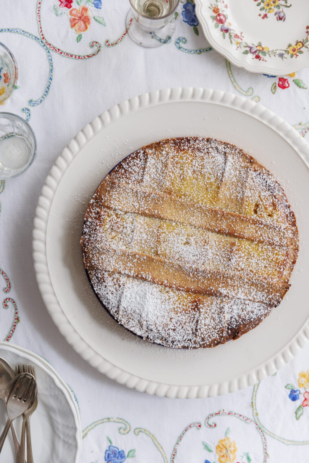 A round cake dusted with powdered sugar on a white platter, set on a table with a white embroidered tablecloth and floral-patterned dishes.