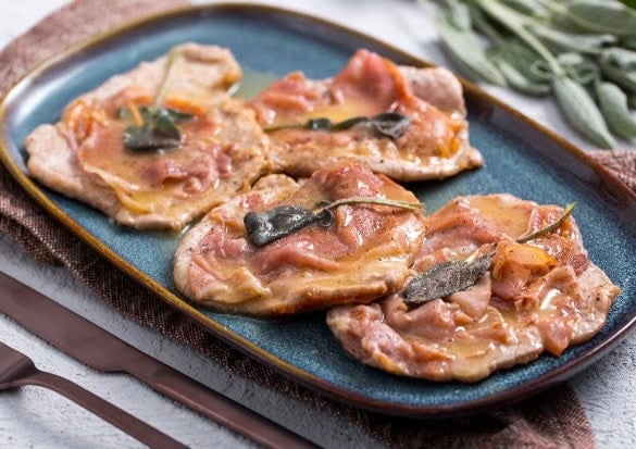 Four pieces of veal saltimbocca topped with prosciutto and sage on a blue oval plate.