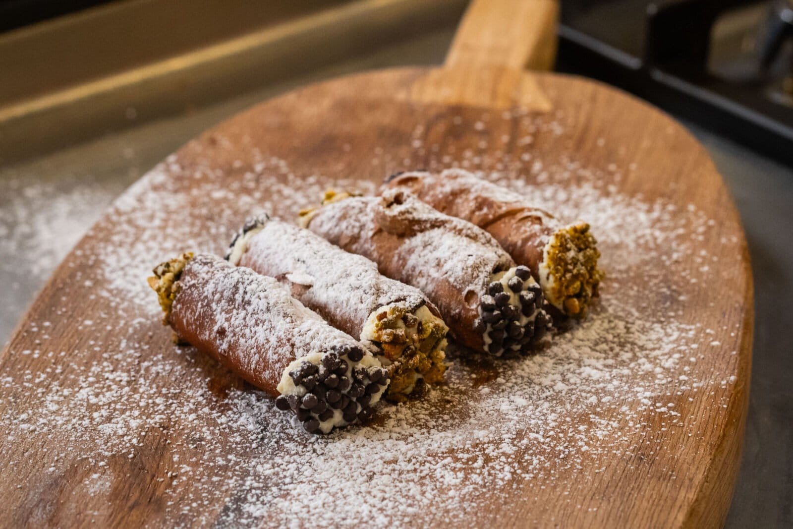 Three cannoli dusted with powdered sugar on a wooden board, with chocolate chips and crushed nuts at one end.