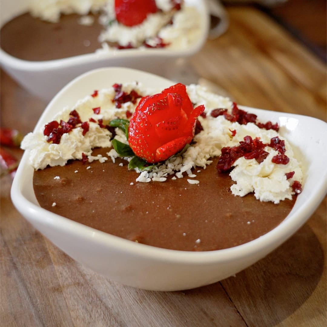 Heart-shaped bowl of chocolate mousse topped with whipped cream, a red strawberry garnish, shredded coconut, and chopped red fruit pieces on a wooden surface.
