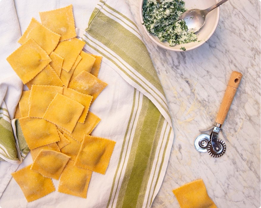 Fresh, uncooked ravioli rest on a striped towel next to a bowl of spinach ricotta filling and a pasta cutter on a marble countertop.