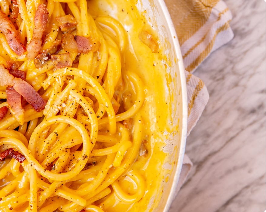 Close-up of spaghetti carbonara in a creamy yellow sauce with pieces of bacon, served in a white dish on a marble surface.