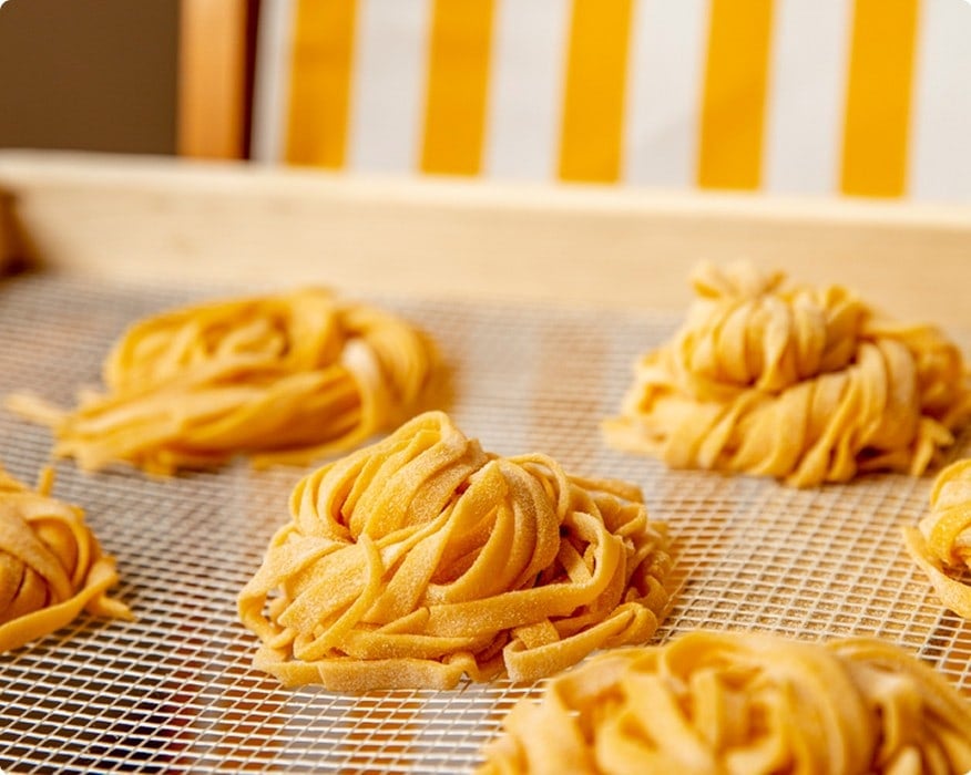 Fresh nests of uncooked tagliatelle pasta rest on a mesh drying rack with a wooden frame and a yellow-striped background.