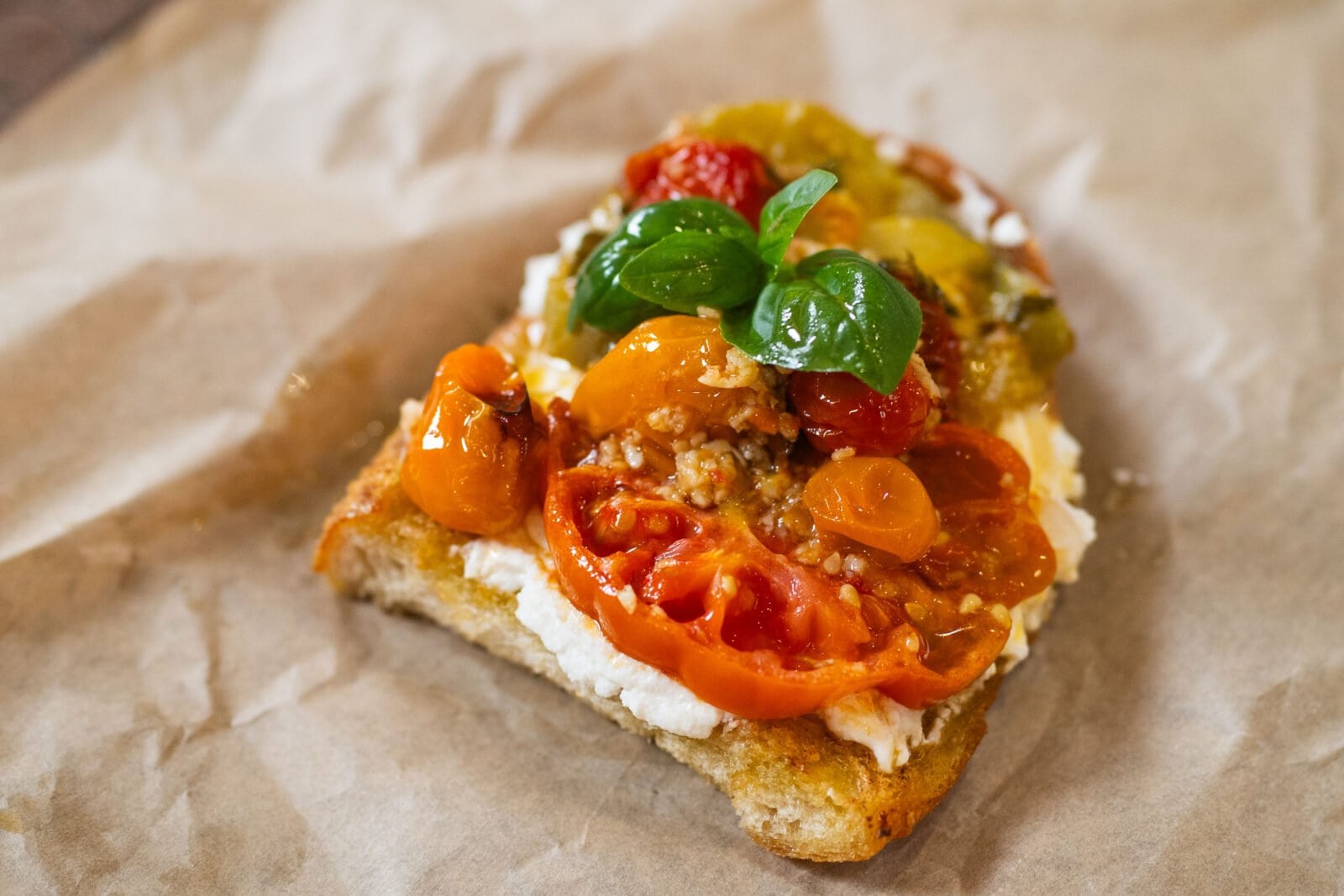 A slice of bread topped with creamy cheese, sliced tomatoes, cherry tomatoes, and fresh basil leaves on parchment paper.