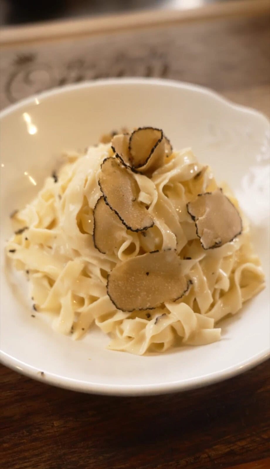 A bowl of fettuccine pasta with cream sauce, topped with thin slices of black truffle on a white plate.
