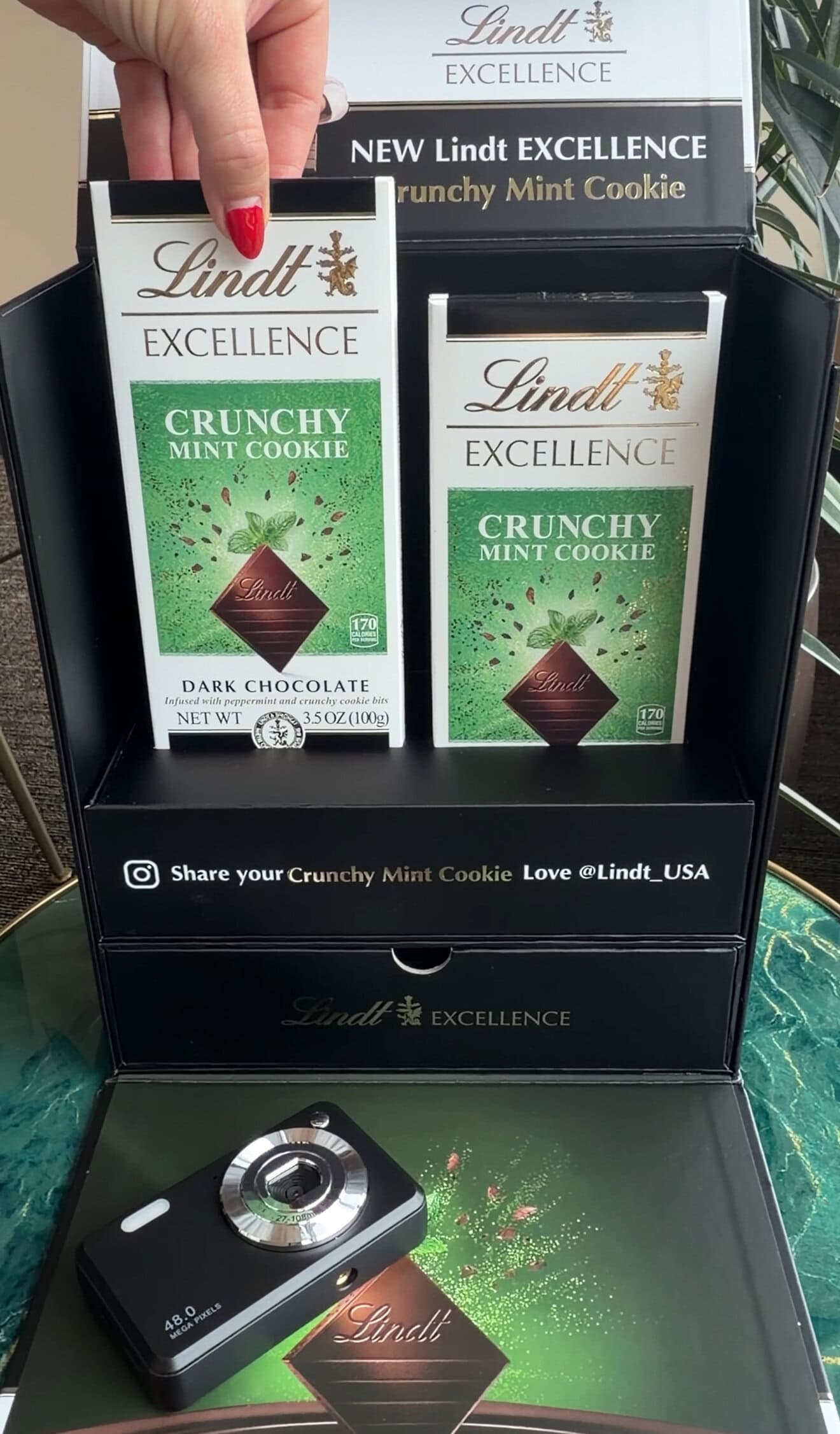 Display of Lindt Excellence Crunchy Mint Cookie dark chocolate bars, with a hand holding one bar, promotional box, and a camera placed in front.