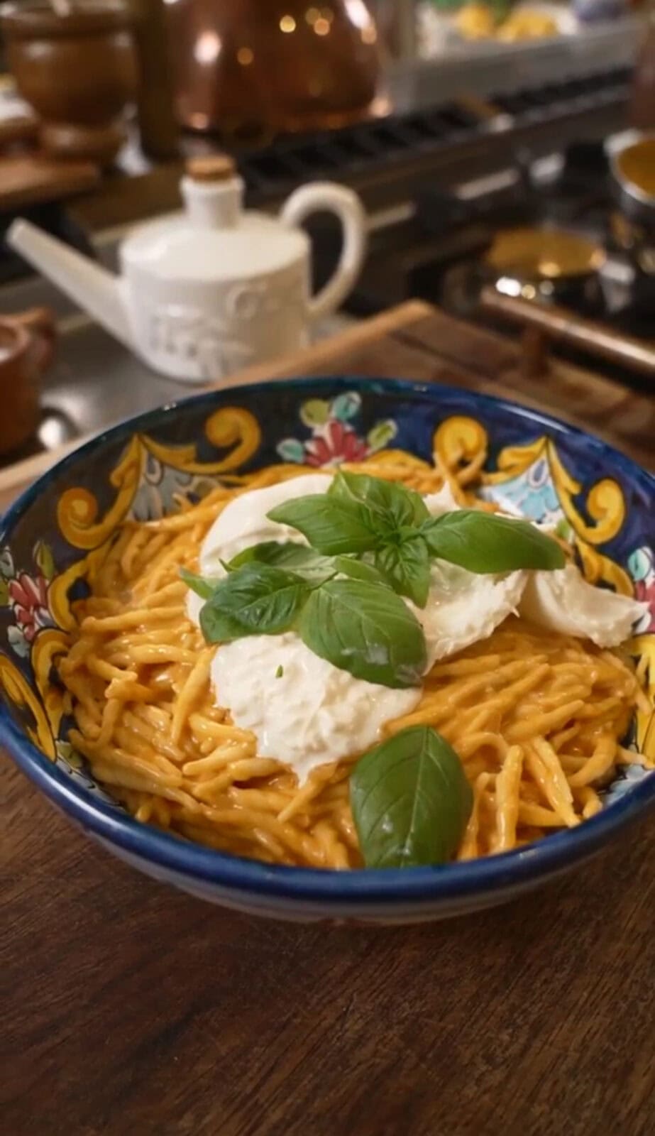 A colorful bowl of spaghetti with red sauce, topped with mozzarella and fresh basil leaves, sits on a wooden counter. A teapot is visible in the background.