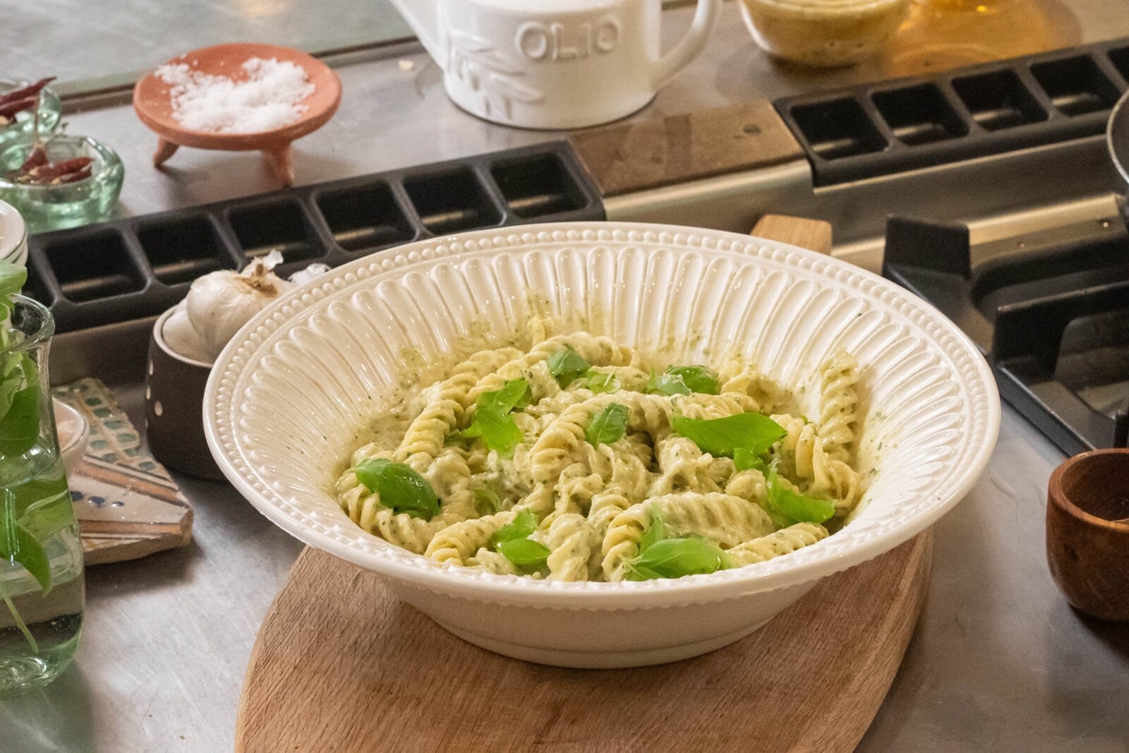 A white bowl of fusilli pasta coated in pesto sauce, garnished with fresh basil leaves, placed on a wooden board on a metal countertop with assorted kitchen items in the background.