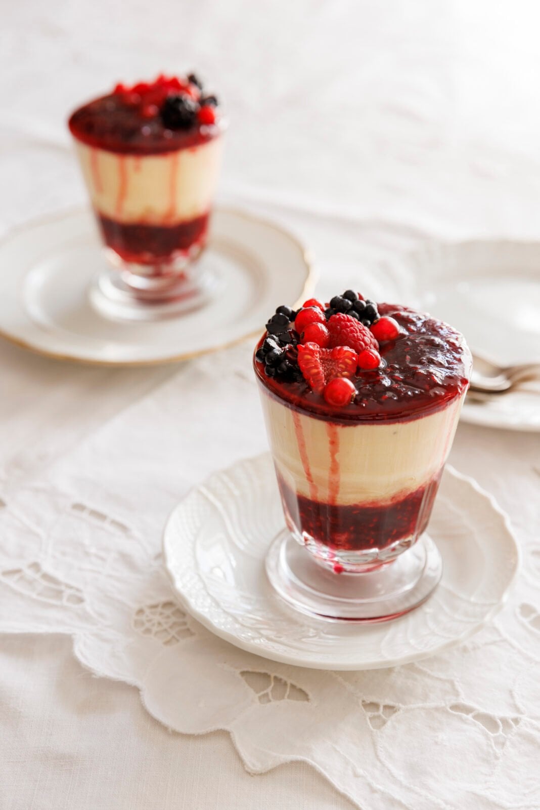 Two glasses of vanilla panna cotta topped with a mixed berry sauce and fresh berries, placed on white plates with linen napkins.