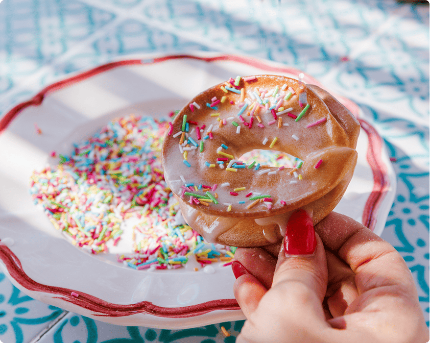 A hand with red nails holds a frosted donut with colorful sprinkles; a plate with more sprinkles is visible in the background.