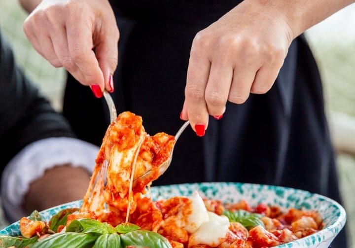 A person serves gnocchi with tomato sauce and basil from a decorative dish onto a plate at a table.