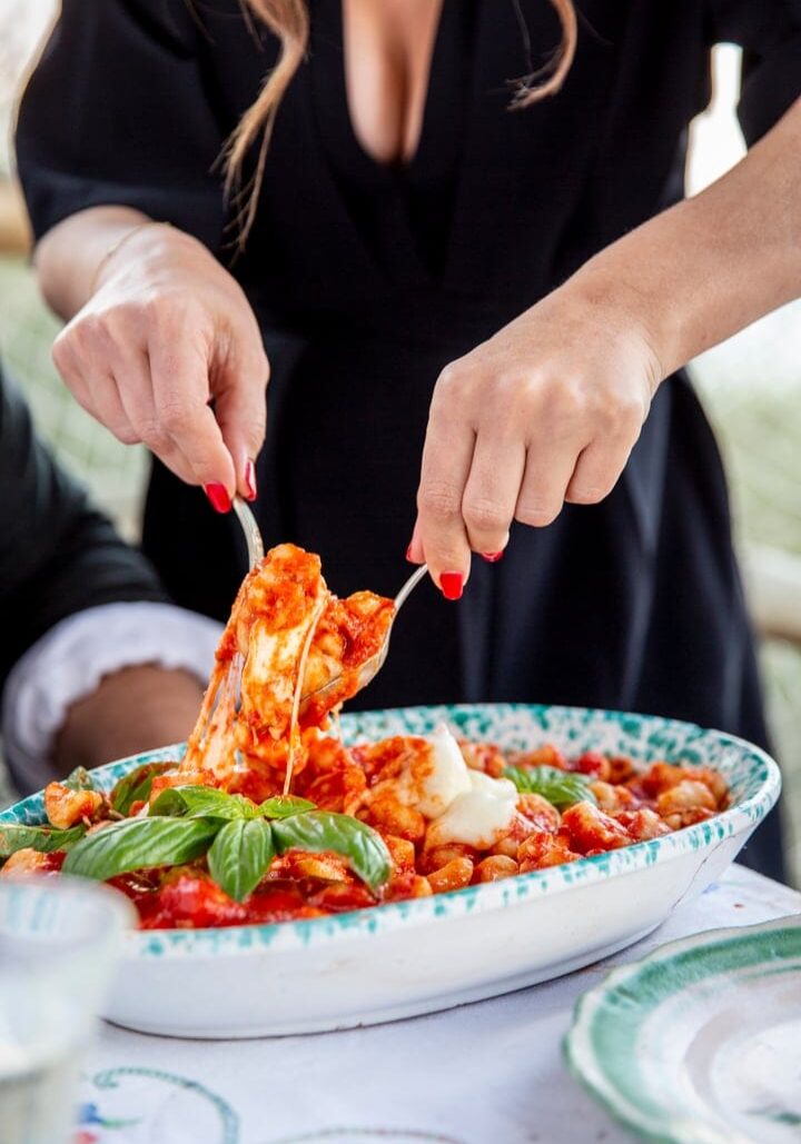 A person serves gnocchi with tomato sauce and basil from a decorative dish onto a plate at a table.