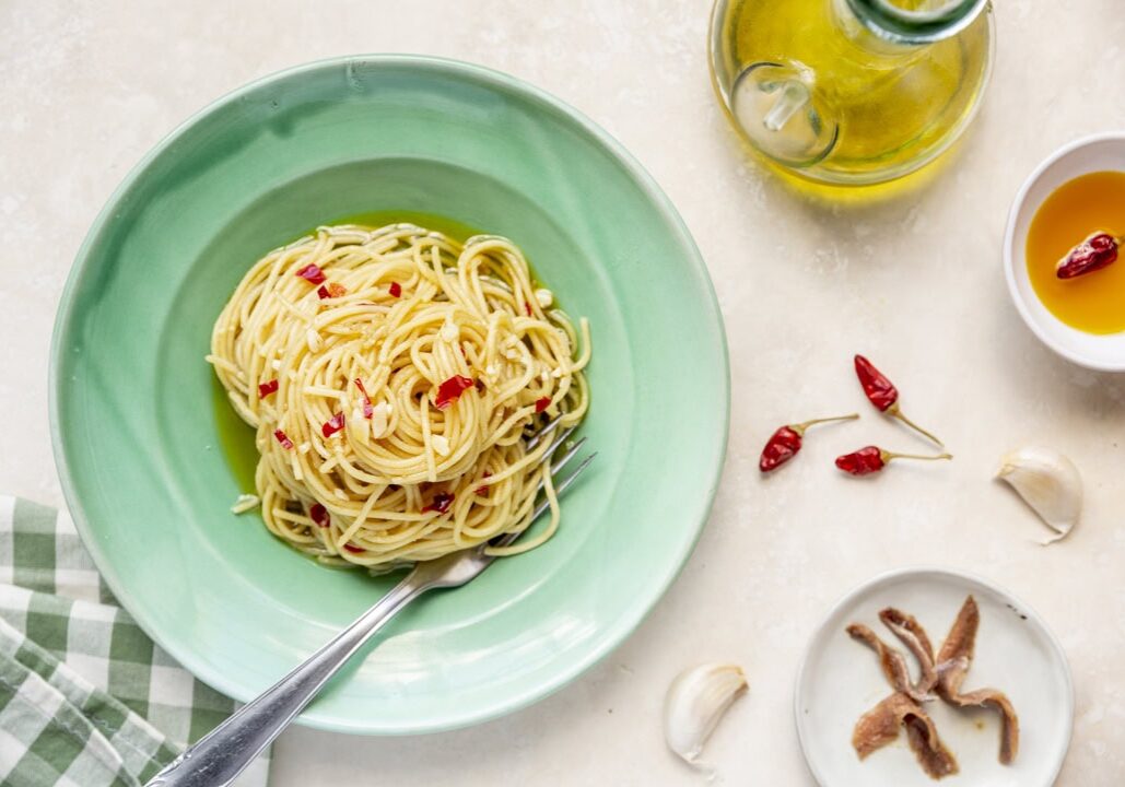 A plate of spaghetti aglio e olio with red chili flakes on a green plate. Nearby are a bottle of olive oil, garlic cloves, a bowl of oil with chilies, and anchovies on a small plate.
