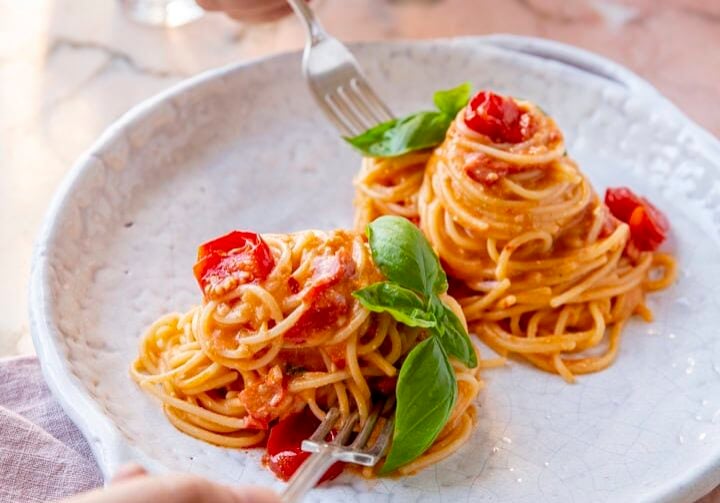 Two people use forks to eat spaghetti with tomato sauce and basil on a white plate, set on a marble table with a glass of water in the background.