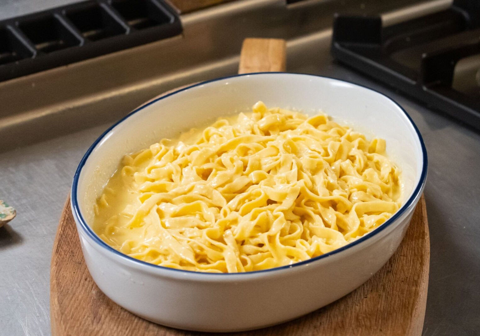 A white oval dish filled with cooked fettuccine pasta coated in a creamy sauce sits on a wooden board next to a stovetop.