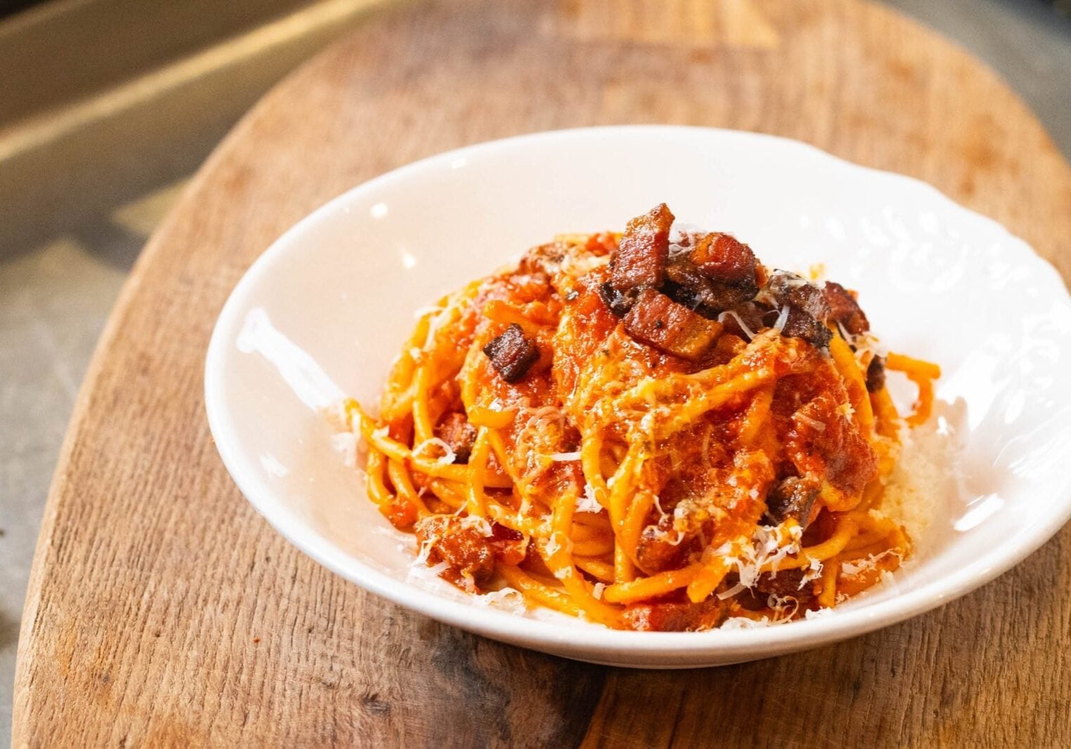 A white bowl filled with spaghetti topped with tomato sauce, pieces of meat, and grated cheese on a wooden surface.
