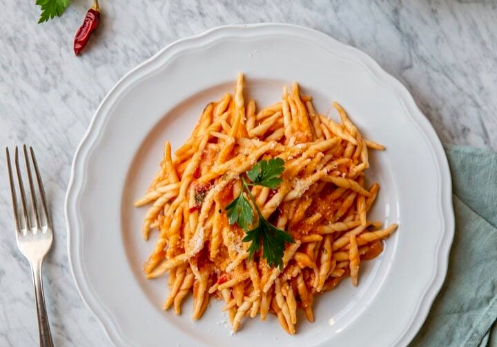 A plate of pasta with tomato sauce and a parsley garnish on a marble surface, accompanied by a fork, chili peppers, and a cloth napkin.