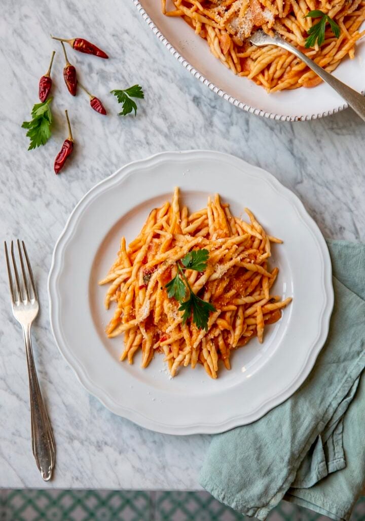 A plate of pasta with tomato sauce and a parsley garnish on a marble surface, accompanied by a fork, chili peppers, and a cloth napkin.