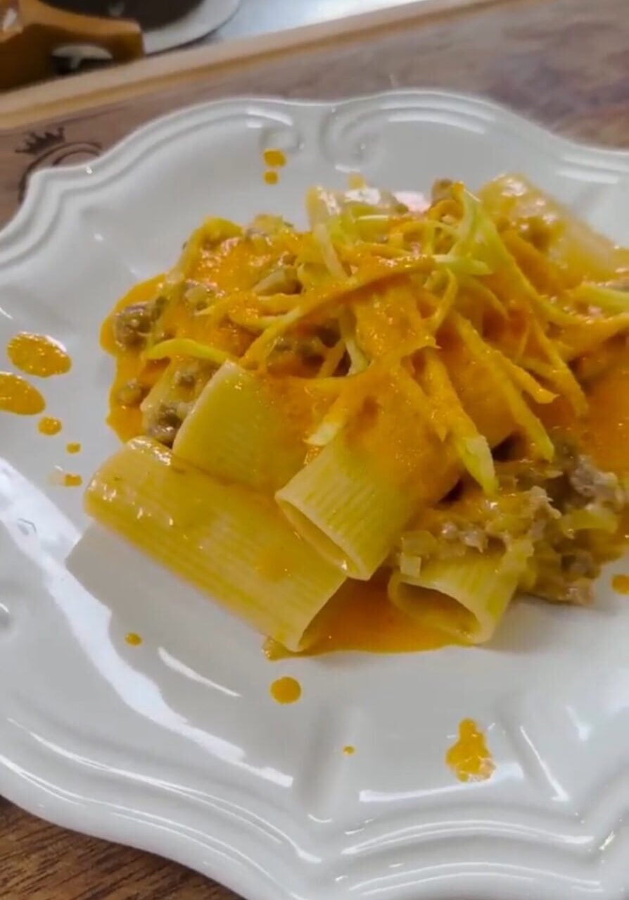 A white plate with rigatoni pasta topped with a rich orange sauce and shredded cheese, garnished with small pieces of meat.